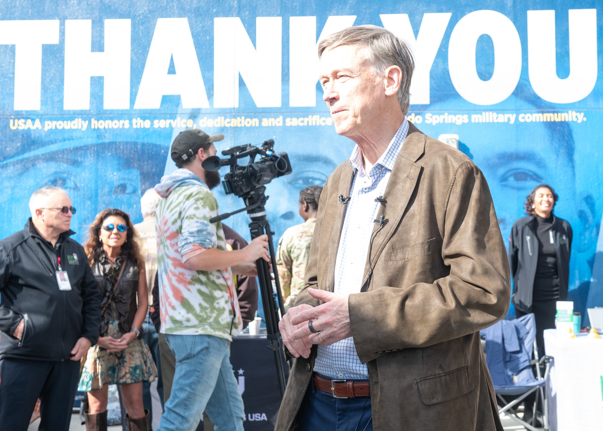 A senator in a brown blazer speaking at an outdoor event, with a large 'THANK YOU' sign in the background and people, including a camera crew, gathered behind him.