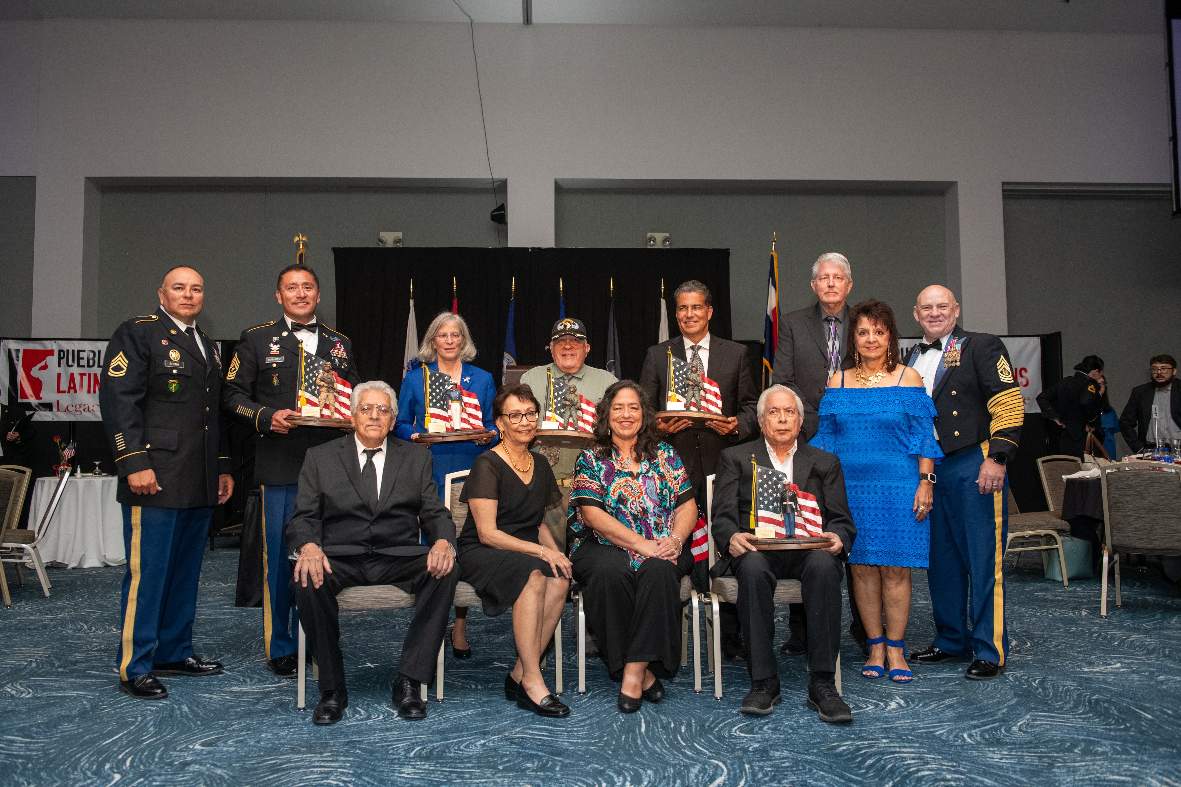 Group photo of attendees at the 9th Annual Pueblo Latino Veterans 'Legacies of Courage' event, featuring men and women in formal attire and military uniforms, some holding awards, posed in front of a stage with flags in the background.