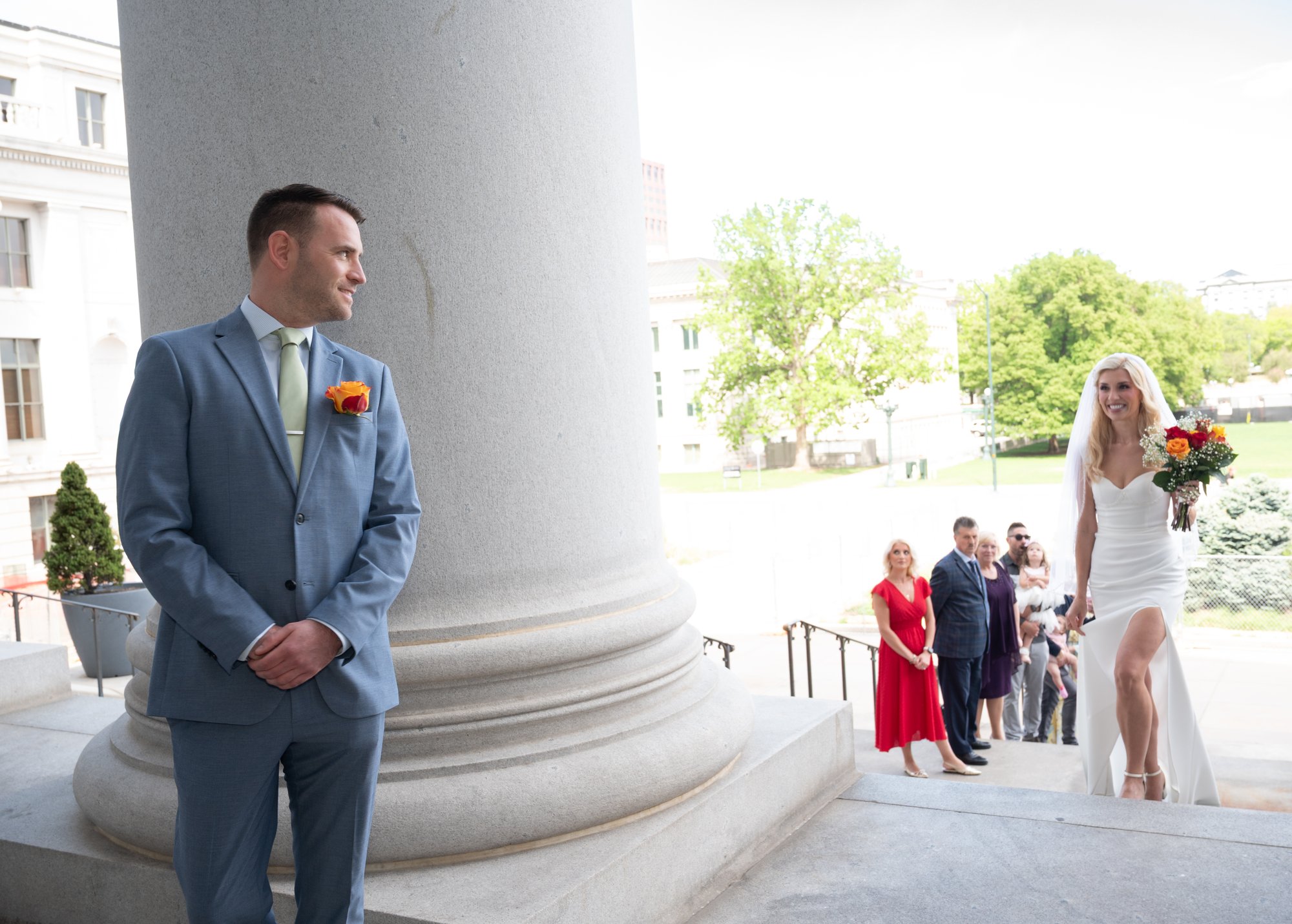 Bride and groom stand before classical columns in a formal wedding portrait, the architectural grandeur framing their refined elegance and the beginning of their enduring legacy.
