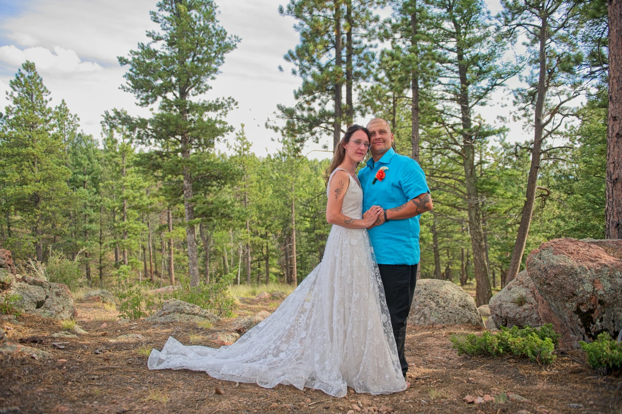 Bride and groom embracing outdoors in a forest setting, with the bride wearing a lace wedding gown and the groom in a blue shirt, surrounded by pine trees and natural rock formations.