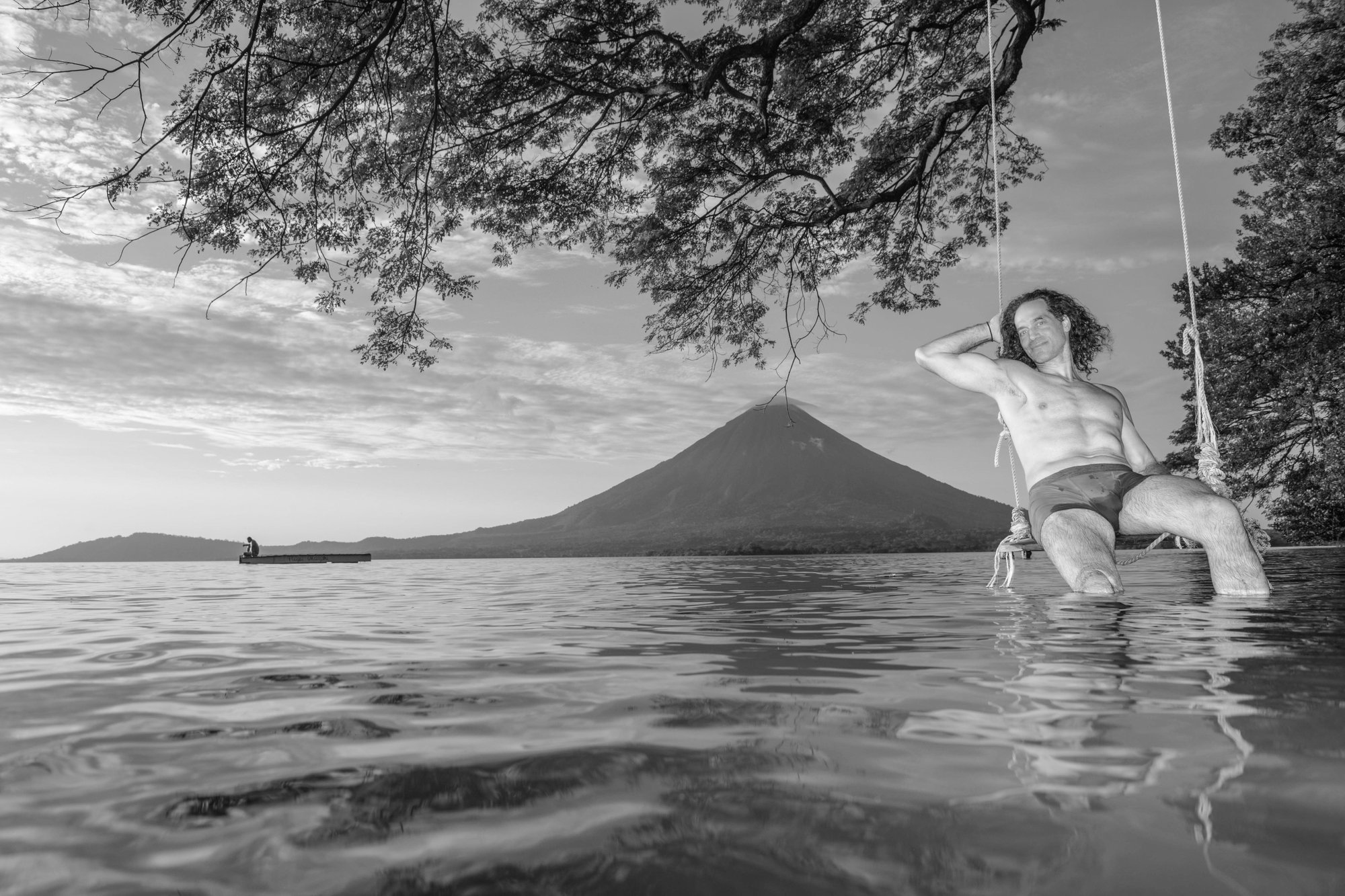 Black-and-white photo of a woman standing in shallow water at Lake Nicaragua on Ometepe Island, posing beneath a tree with a volcanic mountain rising in the background.