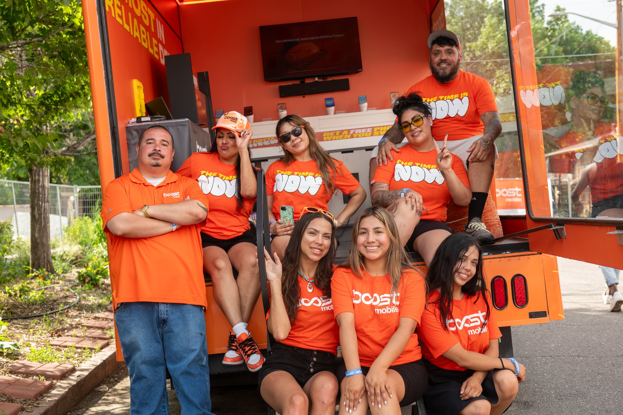 Group of smiling people wearing matching orange shirts posing together in and around an orange food truck, outdoors on a sunny day.