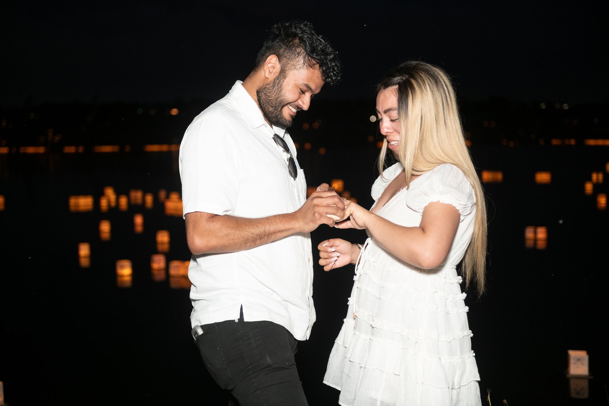 Couple standing together at night during a romantic proposal, with the man placing a ring on the woman’s finger as candlelight glows in the background.