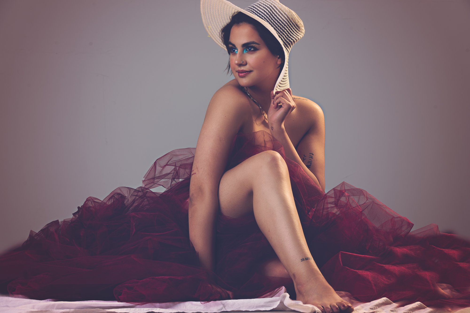 Studio portrait of a woman seated on fabric, wearing a wide-brim hat and an elegant flowing gown, posing thoughtfully against a soft, neutral background.