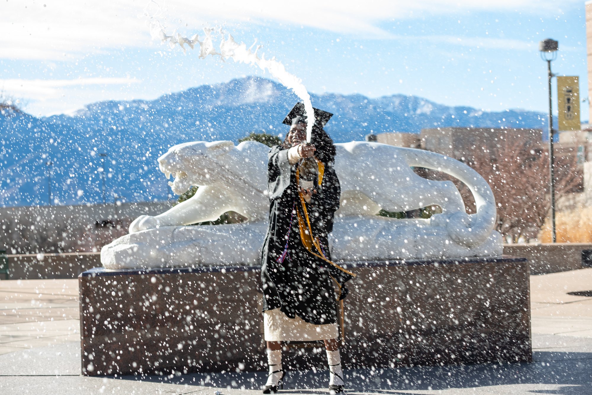 Graduate celebrating outdoors by spraying champagne into the air in falling snow, standing in front of a large white horse sculpture with mountains in the background.