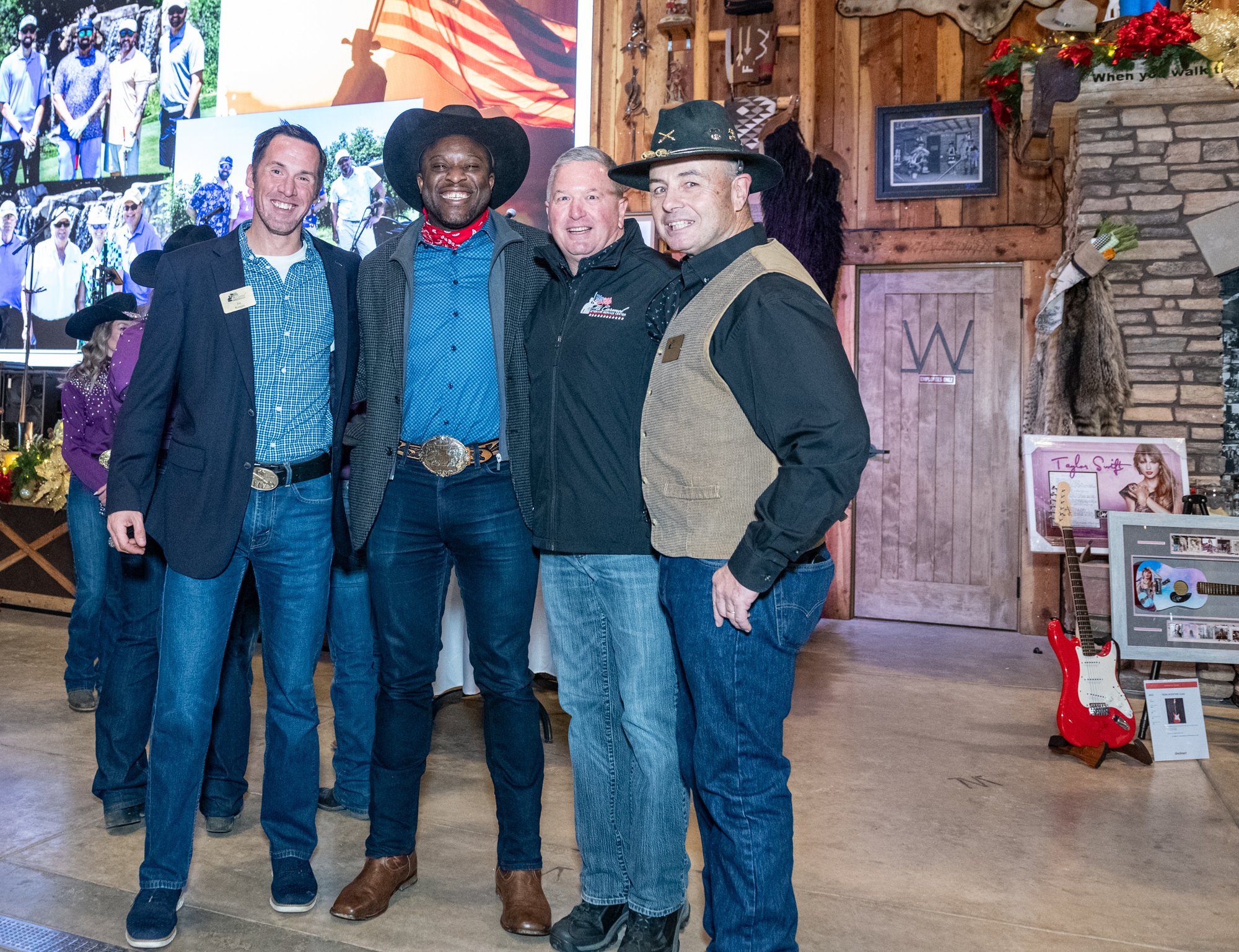 Group photo featuring the current Colorado Springs Mayor, dressed in a blue shirt and cowboy hat, standing with three other men in formal and Western-style attire at an indoor event with wooden decor and a projected image in the background.