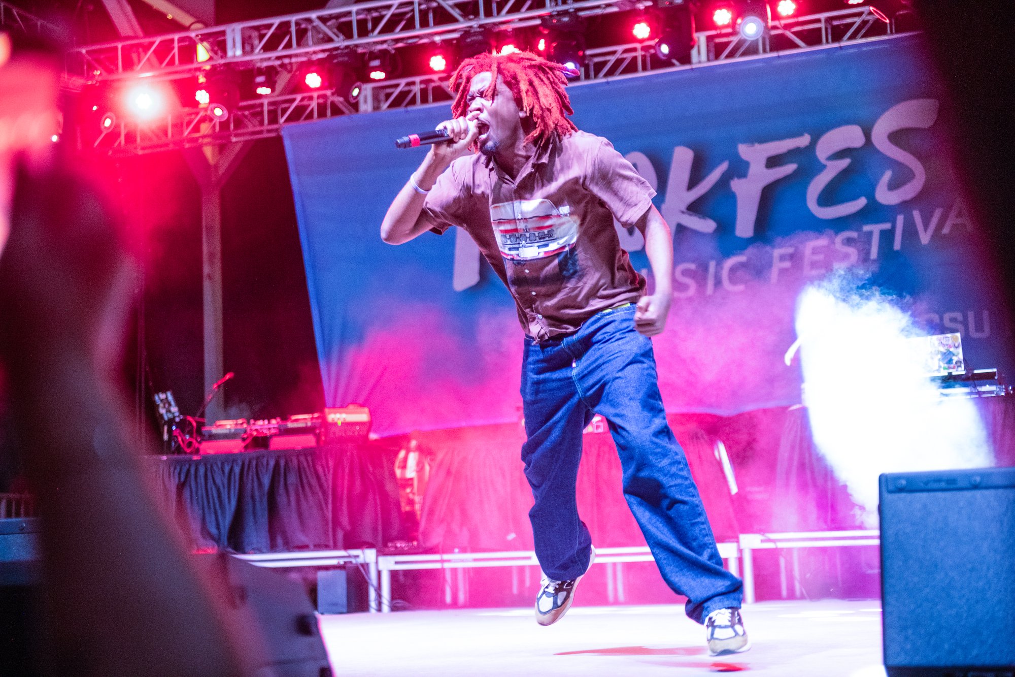 Musical performer singing energetically on stage under red concert lighting, wearing casual streetwear, with a live music festival banner visible in the background.