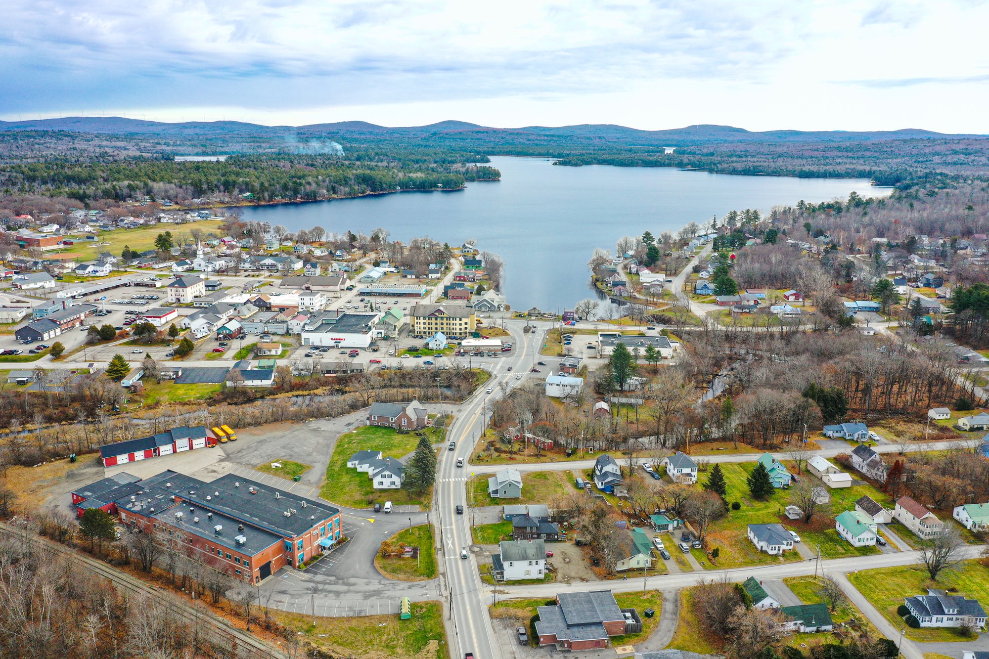 Aerial drone view of the Pueblo Chile Festival park, showing a large red chile pepper installation suspended over green grass near a canal, with people gathered below and city streets and traffic surrounding the park.