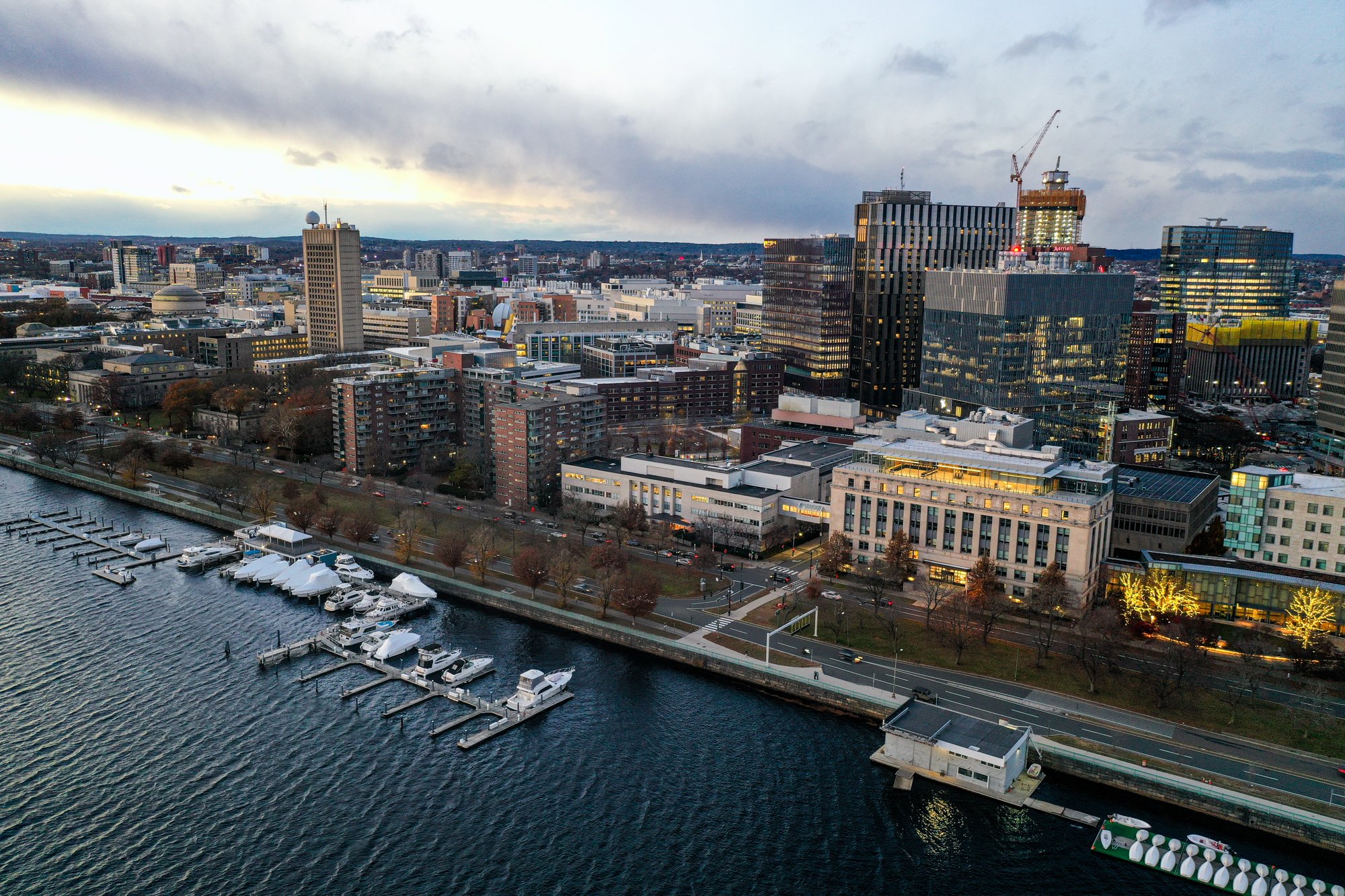Aerial view of Cambridge, Massachusetts, showing the Charles River waterfront, docked boats, and the city skyline with buildings and bridges at dusk. and MIT