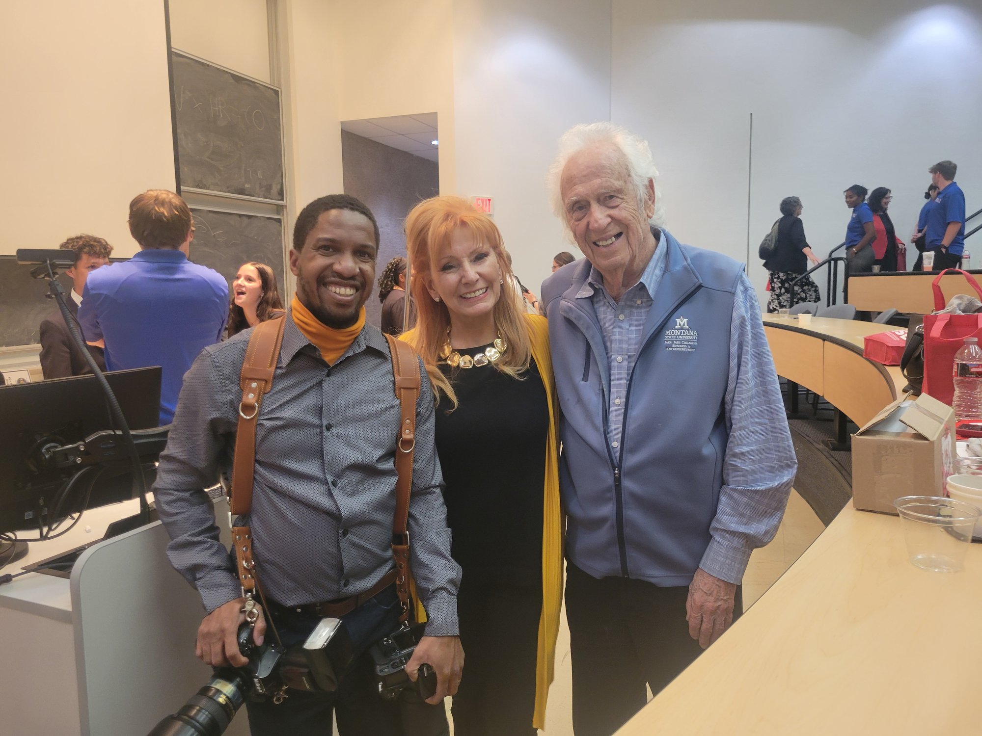 Three people smiling and posing together indoors at a conference or event, including two older adults and a photographer wearing camera gear, with attendees and tables visible in the background.
