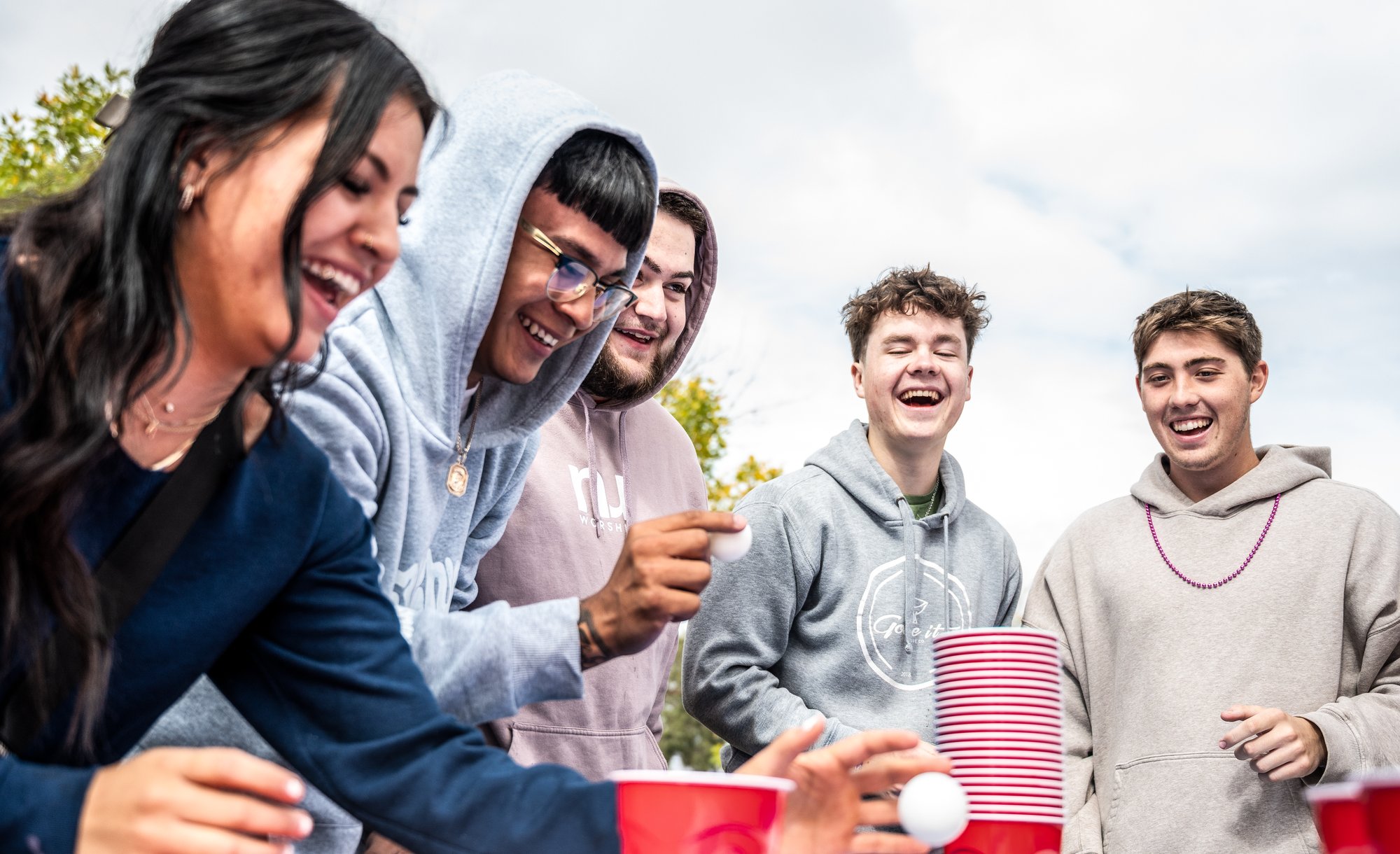 Group of young adults laughing and cheering while playing a cup-stacking party game outdoors, leaning over a table with red plastic cups.