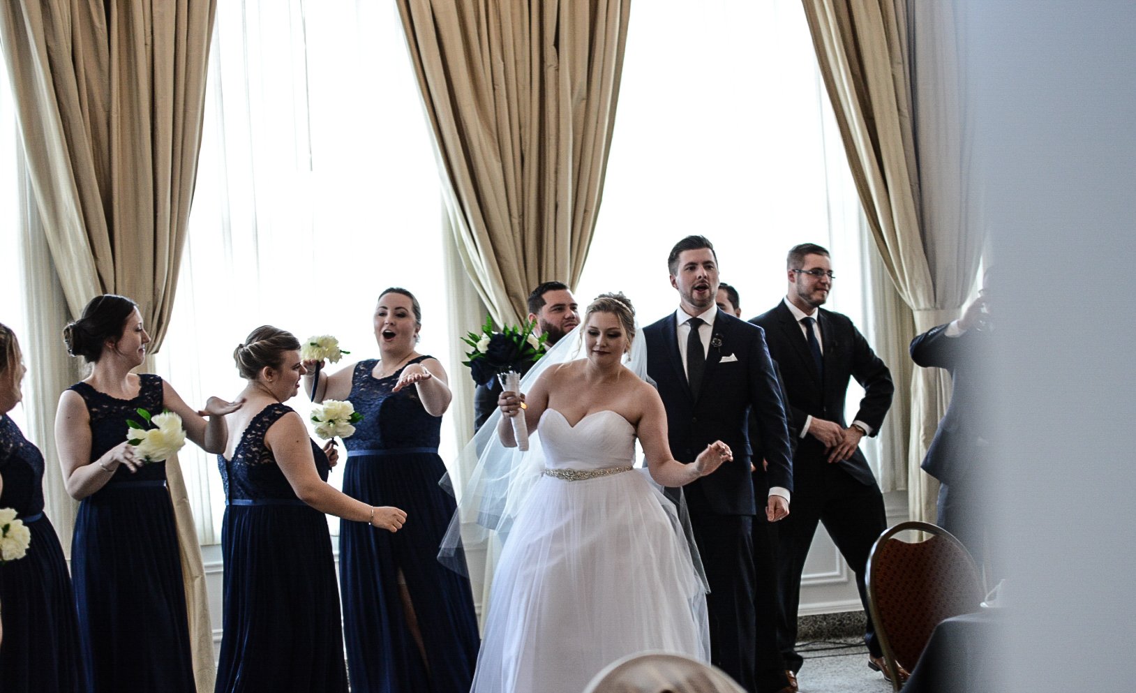 Bride in a white wedding gown walking joyfully with her wedding party indoors, surrounded by bridesmaids in dark dresses and groomsmen in black suits near tall windows.