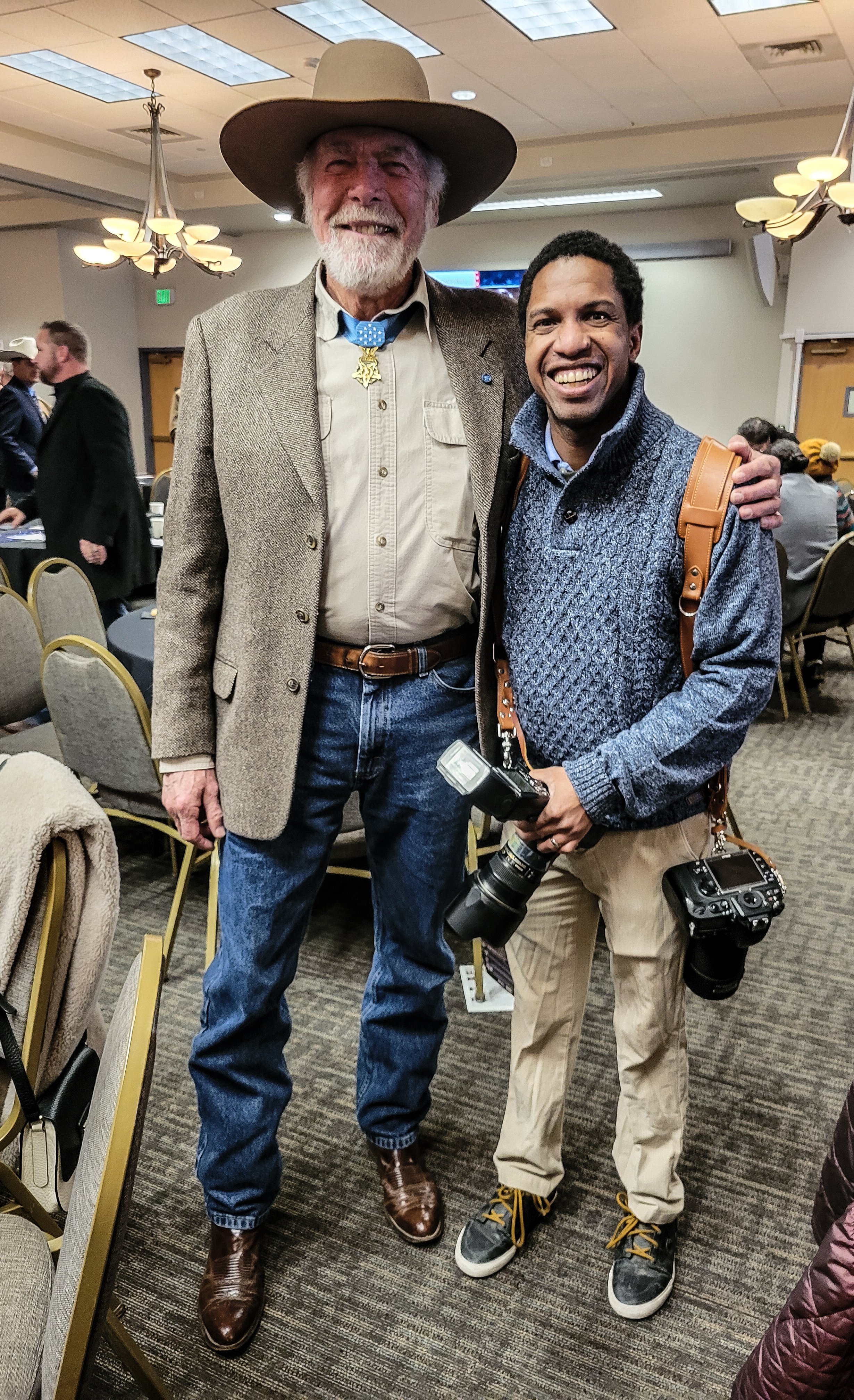 Two men smiling and posing together indoors, one wearing a cowboy hat and Western-style outfit, the other dressed casually with a jacket and camera, standing in a conference or event space.
