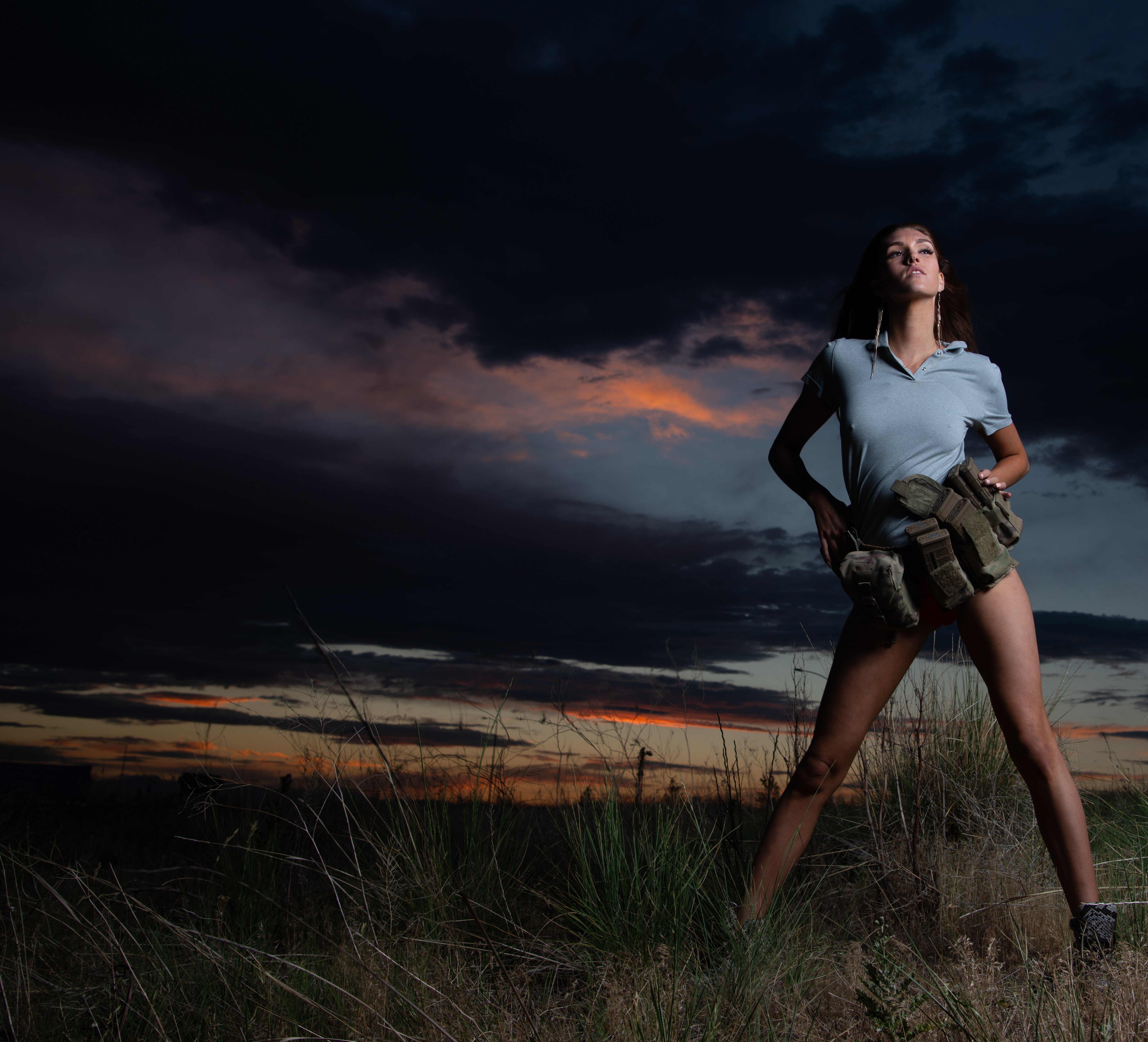 A woman standing confidently in a grassy field during sunset, wearing a light top and shorts, with a dramatic cloudy sky in the background.