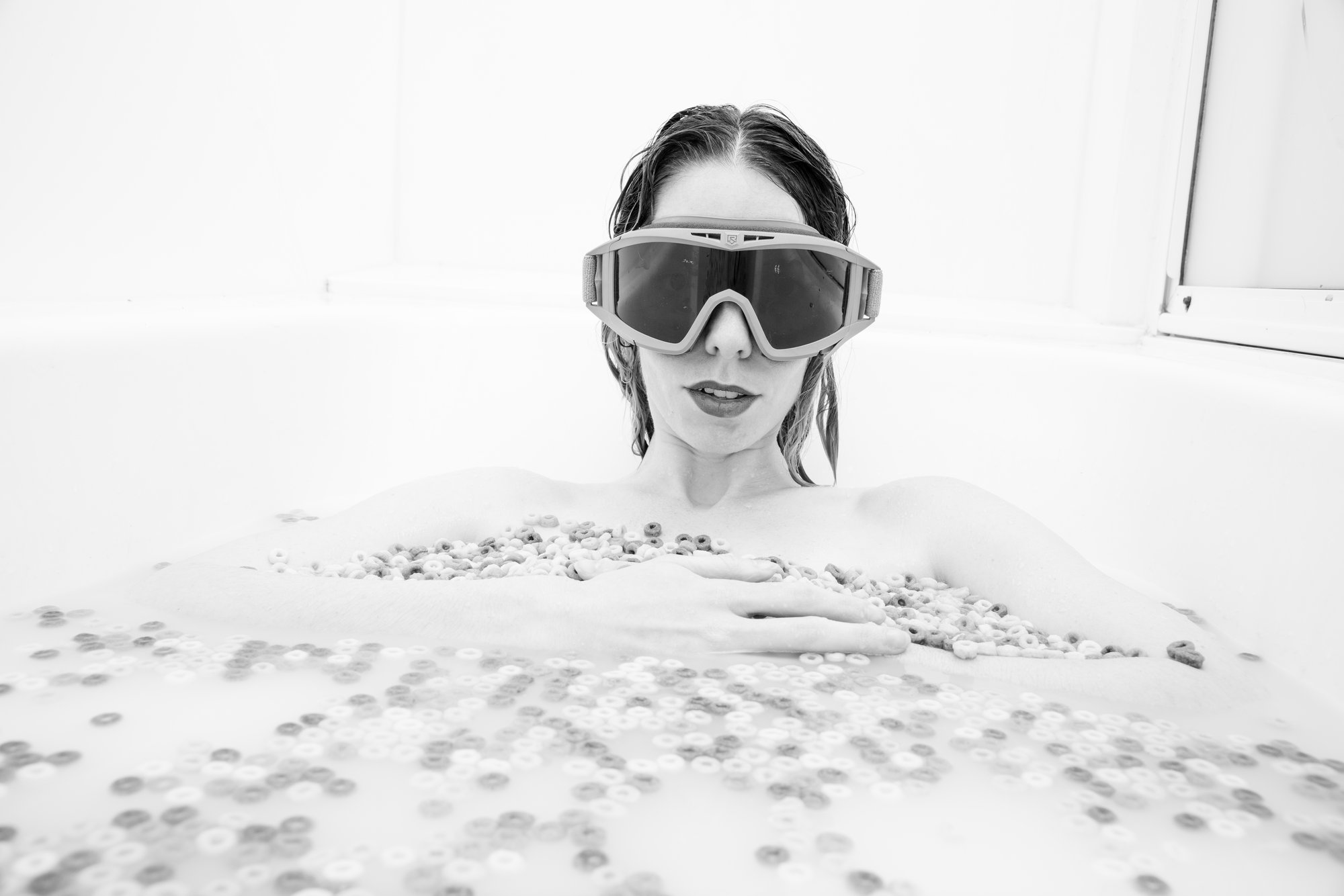 Black-and-white artistic portrait of a woman reclining in a bathtub filled with bubbles, wearing oversized protective goggles, gazing directly at the camera in a playful yet editorial-style setting.