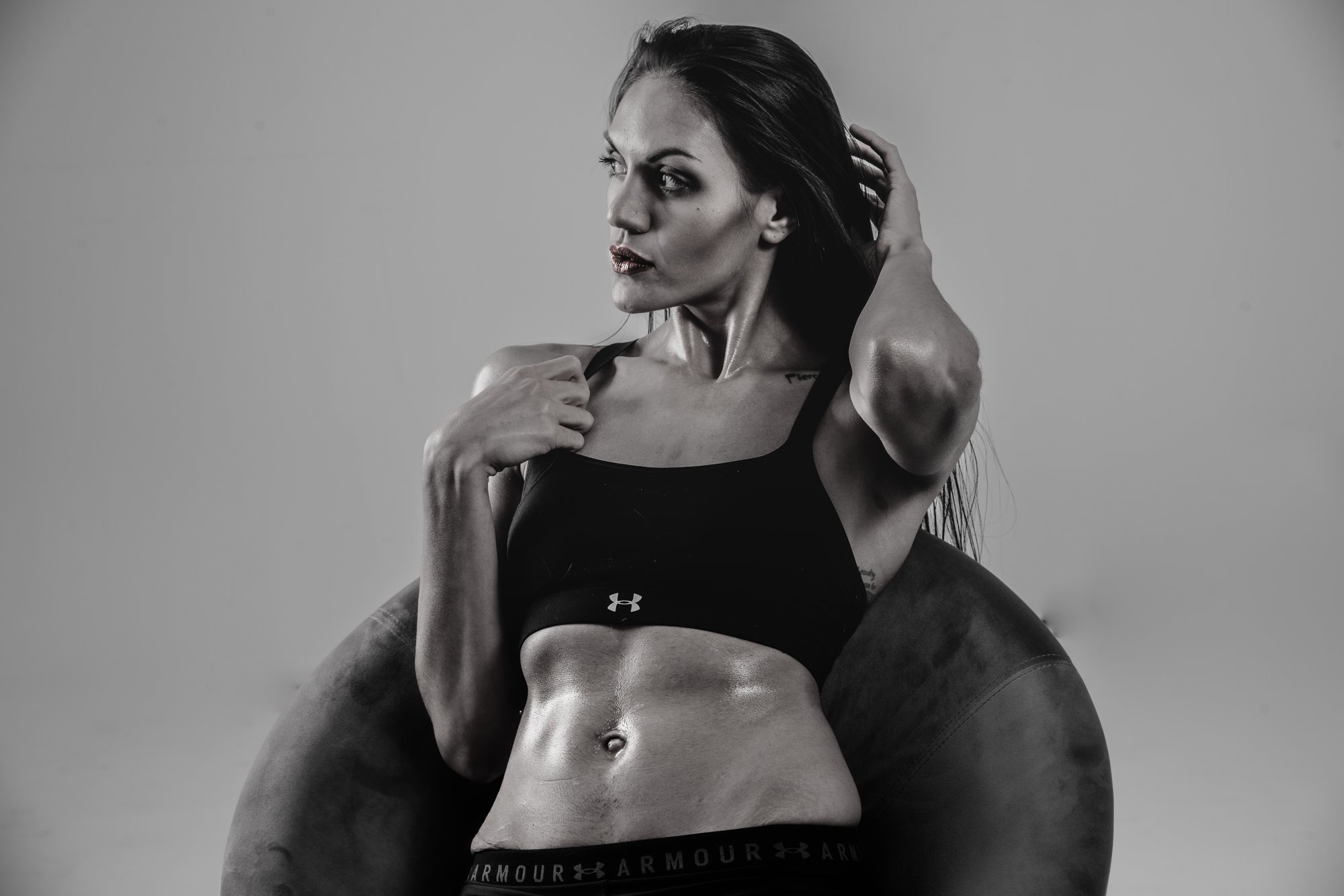 Black-and-white studio portrait of a woman wearing a fitted sports top, posing confidently while seated on a chair, highlighting strength, form, and self-expression against a clean backdrop.