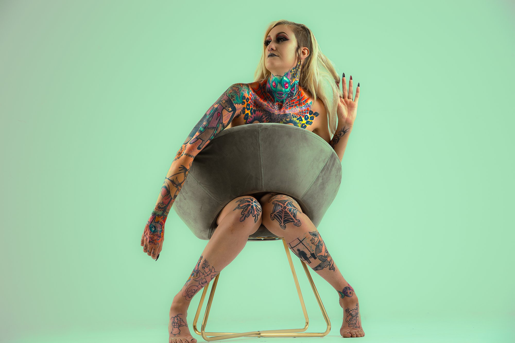 Artistic studio portrait of a tattooed woman seated on a modern chair against a pastel green background, wearing a fashion-forward outfit and posing confidently with expressive body language.