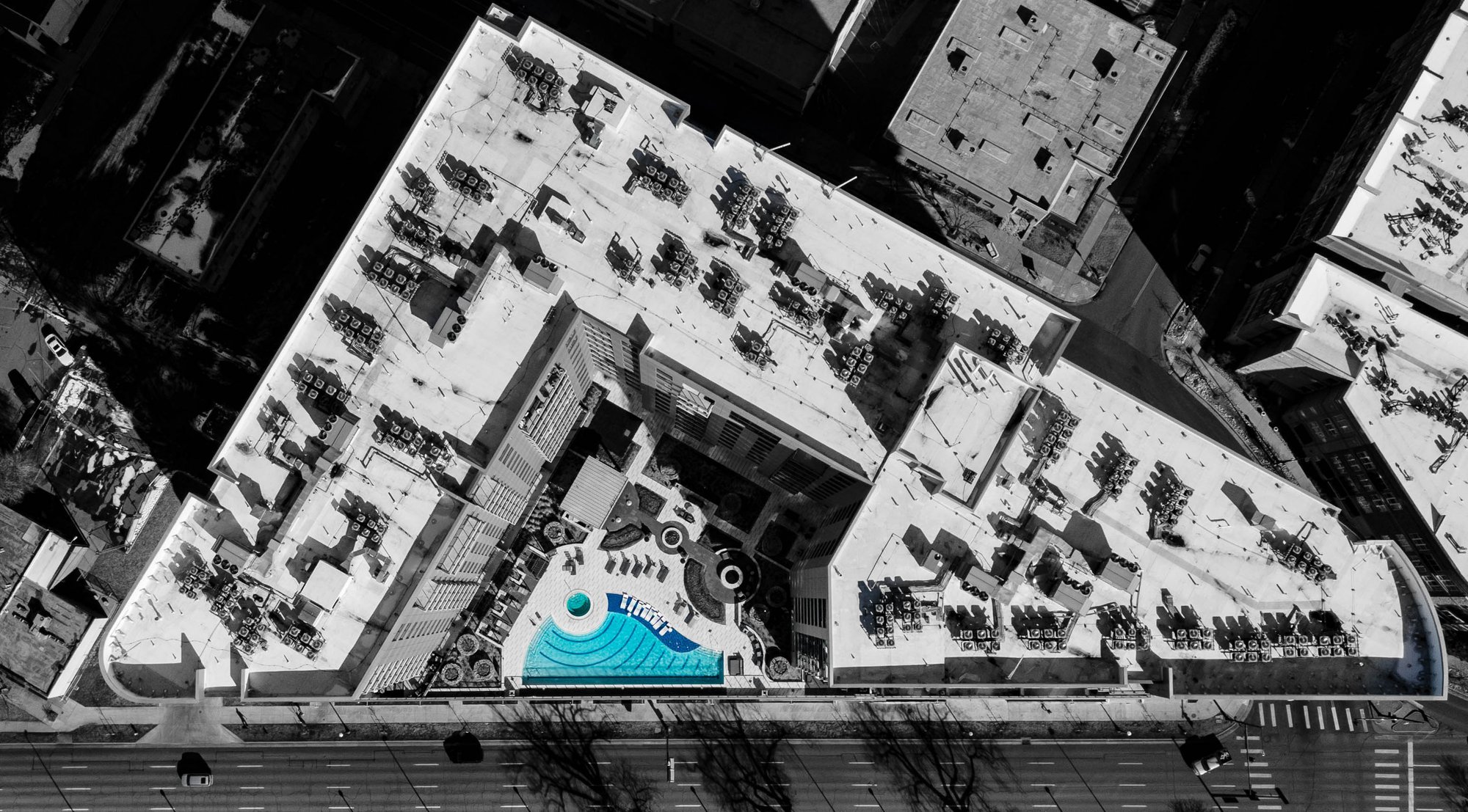 Black-and-white aerial drone view of a famous downtown Denver hotel, showing the building’s geometric rooftop layout with a bright blue outdoor pool visible at the center, surrounded by city streets and neighboring buildings.