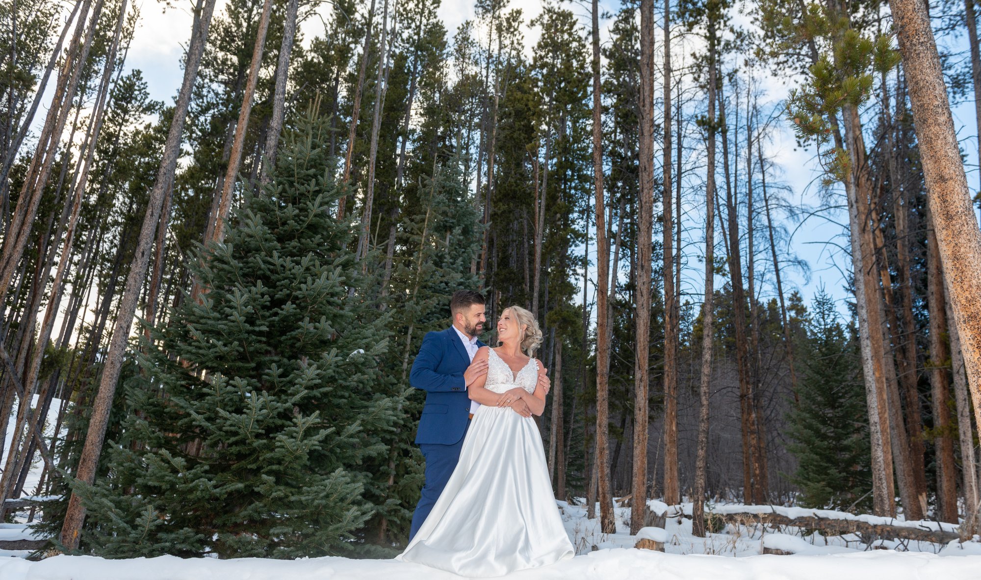 Bride and groom standing close together in a snowy forest, sharing an intimate moment among tall pine trees on a winter wedding day.